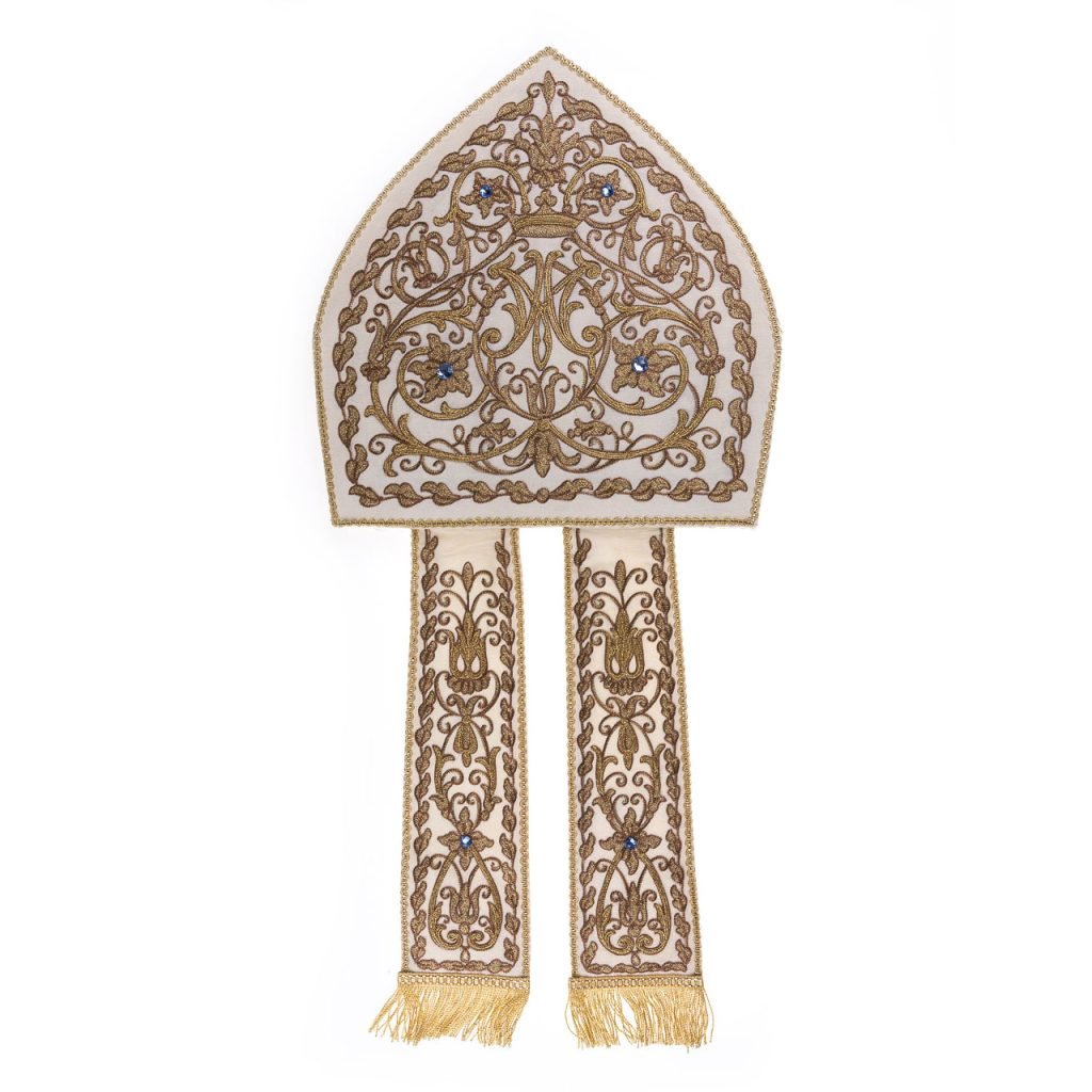 La Mitria AM Ricamata a Macchina è una mitra vescovile in oro e bianco con ricami floreali e lunghe nappe, perfetta per le occasioni cerimoniali.