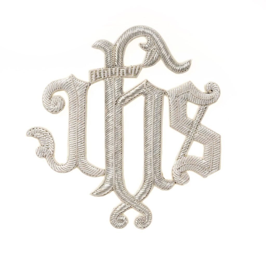 L'Applicazione IHS in Canutiglia presenta un monogramma IHS ricamato in argento con lettere ornate e intrecciate su sfondo bianco.