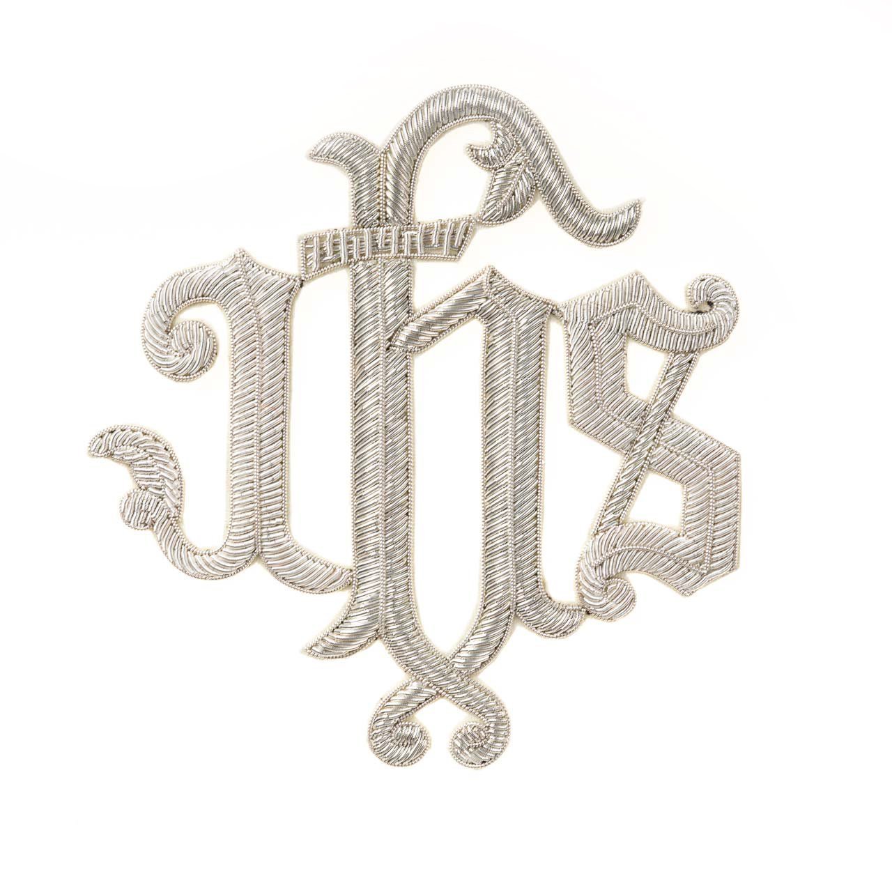 L'Applicazione IHS in Canutiglia presenta un monogramma IHS ricamato in argento con lettere ornate e intrecciate su sfondo bianco.