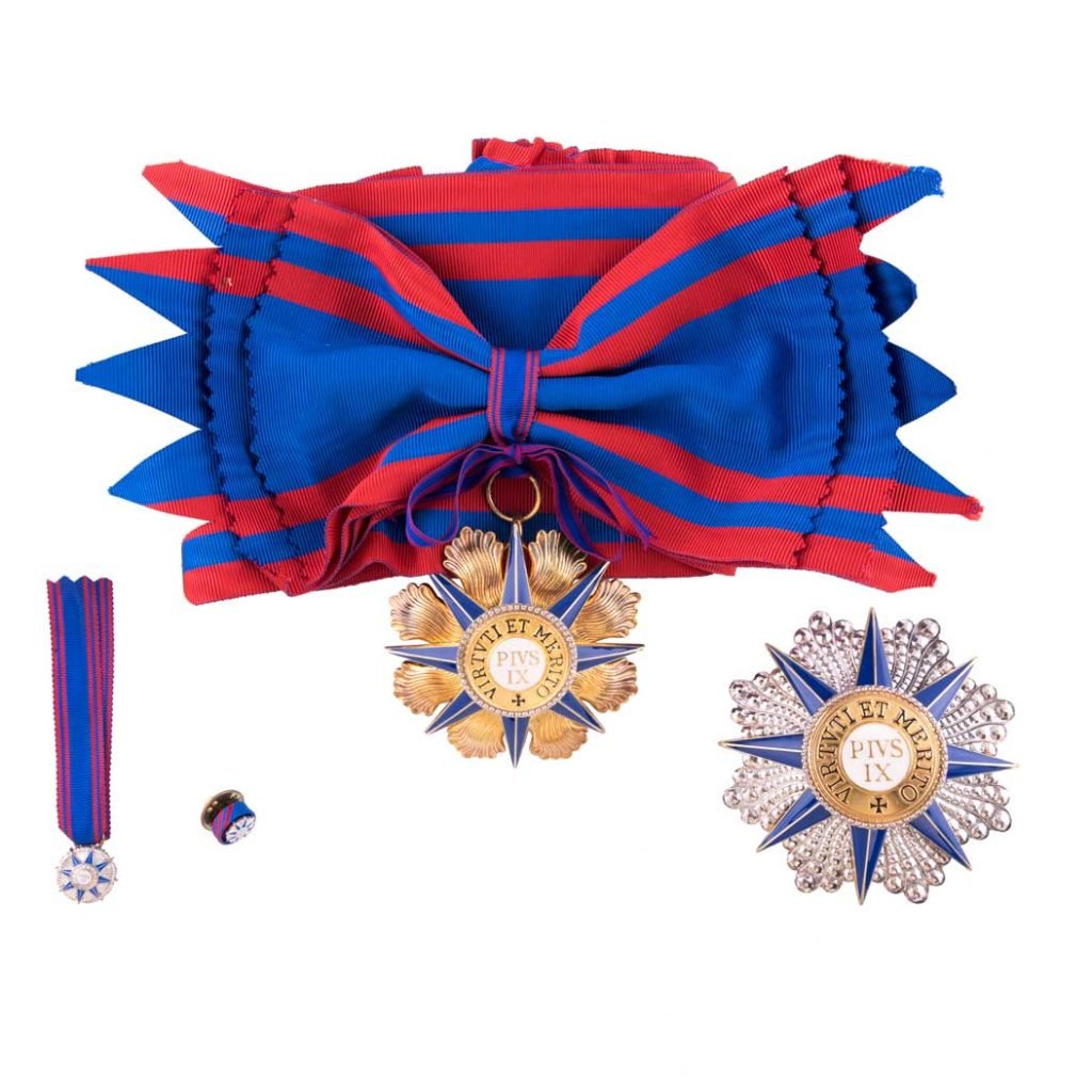 La Decorazione per Cavaliere di Gran Croce Ordine Piano presenta un nastro rosso e blu con medaglie a forma di stella in oro, blu e argento e una spilla, il tutto su sfondo bianco.