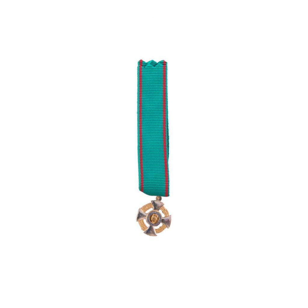 Un nastro verde acqua con bordi rossi trattiene la Decorazione per Cavaliere Ordine al Merito Repubblica Italiana, una medaglia a forma di croce d'oro e d'argento, su sfondo bianco.