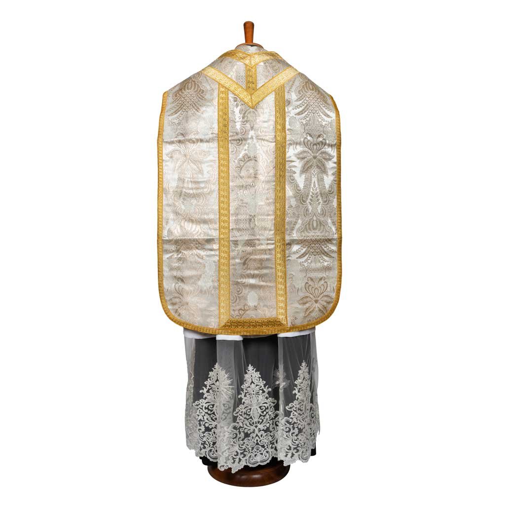 Il Pianete Romana Lampasso GAM1 di Gammarelli Sartoria è un paramento liturgico bianco ornato con finiture dorate e dettagli in pizzo nella parte inferiore, esposto su un manichino.