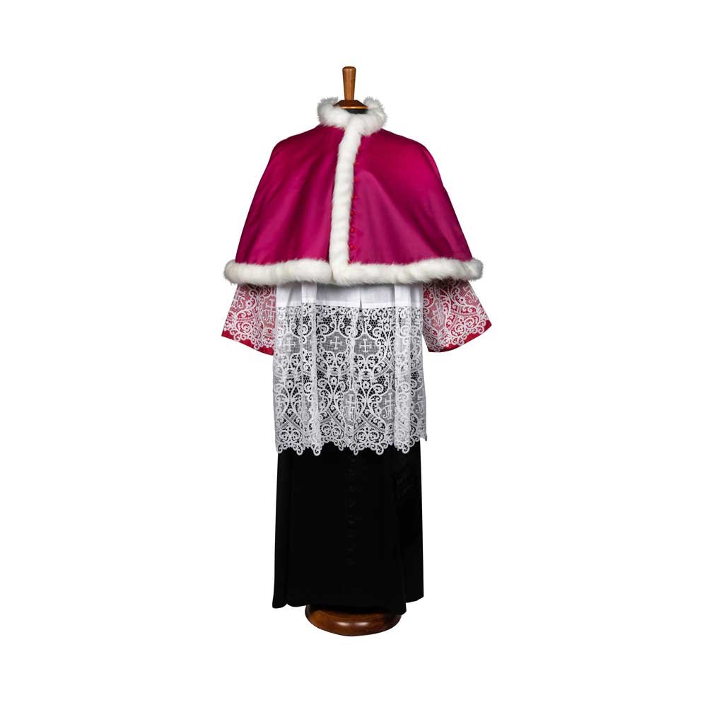 La Mozzetta per Canonici è un paramento clericale con mantello a mozzetta rosa con bordi in pelliccia bianca, maniche in pizzo e una lunga veste nera, esposta su un supporto.