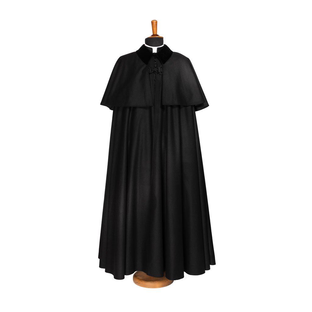 Un Mantò, lungo e nero con un corto mantello, esposto su un supporto di legno su uno sfondo bianco.